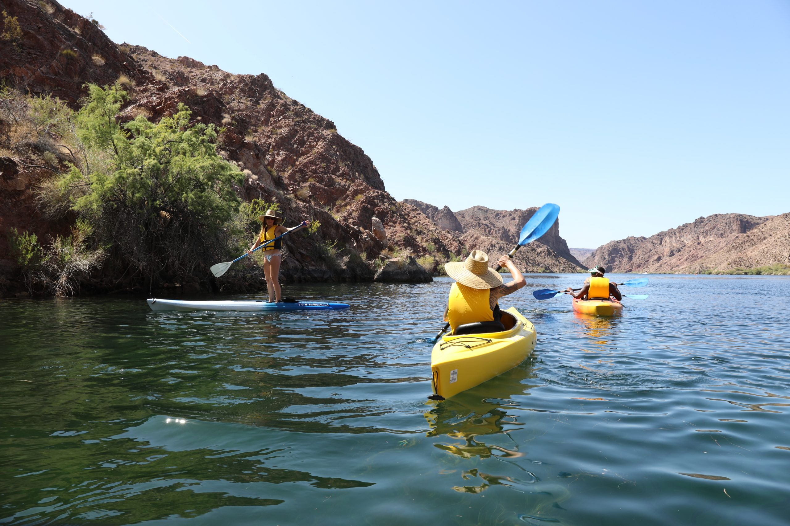 Three people paddle a kayak down a flowing river surrounded by lush greenery and bright blue skies.