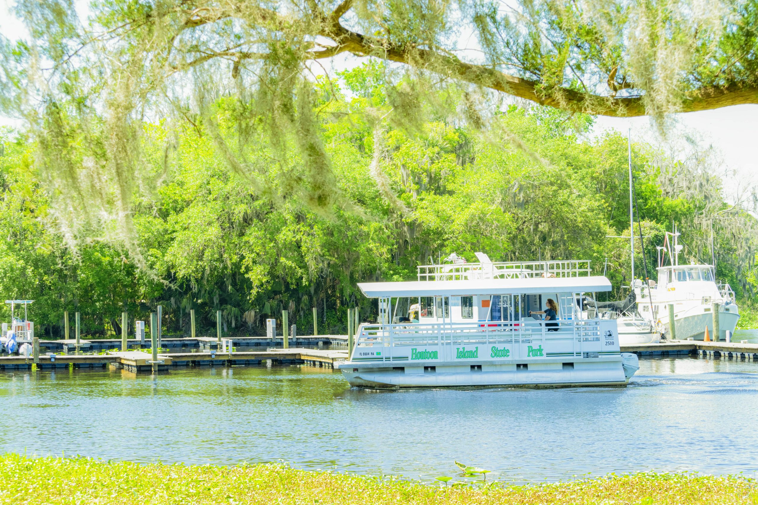 A white and green tour boat cruises along a river, with a marina and lush trees in the background on a bright, sunny day.